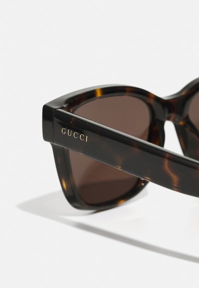 Ochelari de soare din material tartan, cu lentile maro. Rame groase, unghiulare, cu marca "GUCCI" aurie pe laterale. Finisaj neted și lucios.