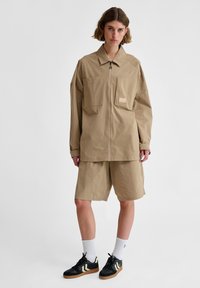 Licht beige utility jack met twee voorzakken, korte mouwen en een rits. In combinatie met bijpassende shorts en zwarte sneakers.
