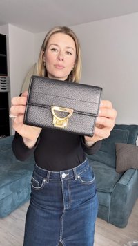 Juodos odos piniginė su tekstūruota paviršiaus faktūra, auksiniu geometriniu užsegimu ir horizontaliomis siūlėmis per priekį.