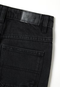 Bolsillo trasero y cinturilla de unos vaqueros de denim negro con una etiqueta de cuero cosida que dice "REPÚBLICA DEL DENIM" sobre un fondo blanco.