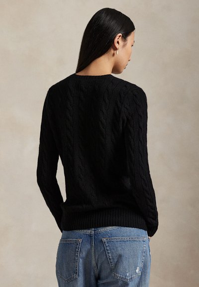 Mujer con cabello largo y oscuro que lleva un suéter negro de punto trenzado y vaqueros azules, vista desde atrás sobre un fondo neutro.