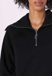 Närbild av en person som bär en svart zip-up huvtröja med en silverdragkedja och en bred krage, som visar den nedre delen av ansiktet och halsen.