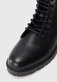 Stivale da combattimento in pelle nera con suola in gomma spessa e lacci neri infilati in occhielli metallici su sfondo bianco.