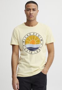 Gul bomulls-T-skjorte med et sirkulært grafisk motiv som viser en sol, bølger, og teksten "BLEND DENIM ESTD 1993". Kort erm og rund hals-design.