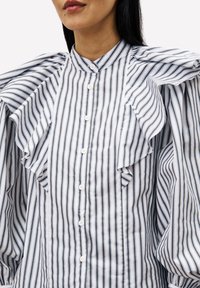 Vrouw in een wit en marineblauw verticaal gestreept blouse met ruffles en witte knopen, weergegeven van nek tot taille.