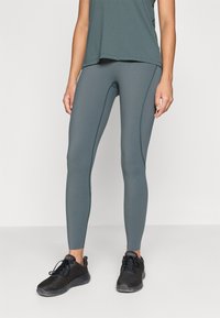 Magas derekú, szűk leggings, tompított zöld színben, kontrasztos varrásokkal. Fekete hálós sportcipővel párosítva.