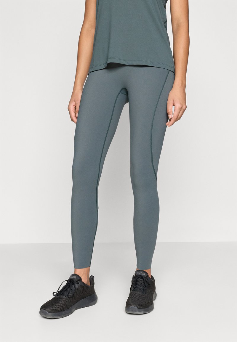 Magas derekú, szűk leggings, tompított zöld színben, kontrasztos varrásokkal. Fekete hálós sportcipővel párosítva.