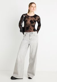 Μαύρο μακρυμάνικο τοπ με δαντέλα και φλοράλ σχέδιο, συνδυασμένο με ανοιχτό γκρι τζιν παντελόνι φαρδιάς γραμμής. Το σύνολο περιλαμβάνει μικρό μαύρο τσαντάκι και χοντρούς μαύρους παπούτσια.