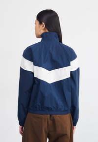 Marineblå vindjakke med et hvidt chevron-design på ryggen, der har høj krave og elastisk kant. Glat, letvægtsstof.