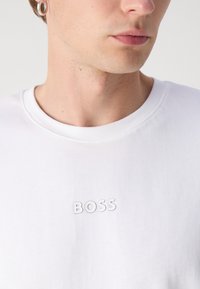 Hvit bomull t-skjorte med ribbestrikket rund hals og en subtil preget "BOSS" logo på forsiden. Glatt tekstur, minimalistisk design.