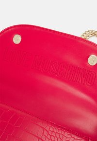 Interiér červené kožené kabelky s raženým logem "LOVE MOSCHINO" a zlatými magnetickými zapínáními připevněnými na zlatém řetízkovém popruhu.