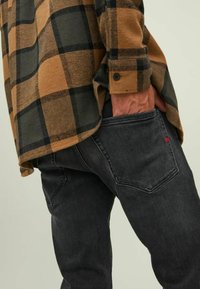Karirano flanelasto srajco v oranžni in črni barvi z gumbnimi manšetami, v kombinaciji s temno sivo denim hlačami, ki imajo rdeč logotip na žepu.