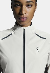 Chaqueta de running blanca ligera con cuello alto, cremallera negra y acentos en negro contrastantes en los hombros. Presenta un logo en el pecho.