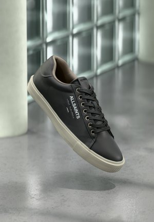 Must AllSaintsi nahast toss, millel on valge tald, mustad paelad ja küljele kinnitatud logo, ujuv halli peegelduva põranda kohal.