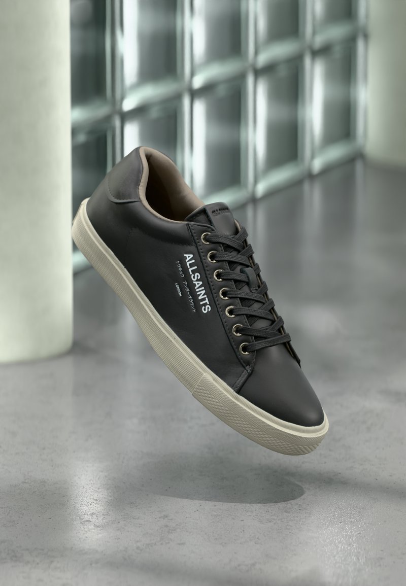 Μαύρο δερμάτινο αθλητικό παπούτσι AllSaints με λευκή σόλα, μαύρα κορδόνια και λογότυπο στο πλάι, αιωρούμενο πάνω από γκρι αντανακλαστικό πάτωμα.