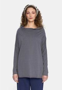 Kobieta z kręconymi włosami, ubrana w szary, długi rękaw, granatowe spodnie i jasnoszary, dziany opaska na głowę, stojąca na jednolitym białym tle.