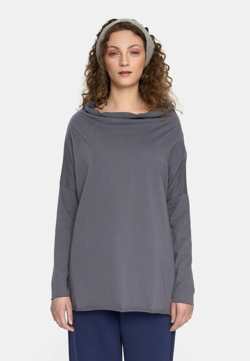 Kobieta z kręconymi włosami, ubrana w szary, długi rękaw, granatowe spodnie i jasnoszary, dziany opaska na głowę, stojąca na jednolitym białym tle.