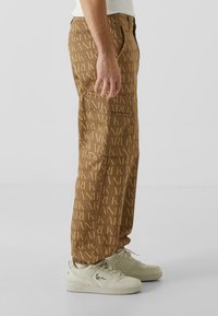 Pantaloni maronii cu un model repetitiv "KARL", talie elastică, buzunare laterale și o croială lejeră. Asortați cu adidași de culoare deschisă.