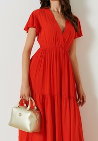 Robe maxi rouge fluide avec de courtes manches volantées et un décolleté en V profond. Tient un sac à main doré brillant avec deux poignées et un fermoir.