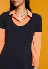 Sukienka z kołnierzykiem, z granatową górną częścią i brzoskwiniowymi długimi rękawami. Dekolt zdobiony falbanami, uzupełniający warstwowy wygląd.