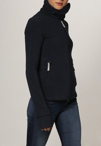 Chaqueta de forro polar azul marino con alto cuello, cierre de cremallera y dos bolsillos frontales. Incluye orificios para los pulgares en las mangas para mayor comodidad.