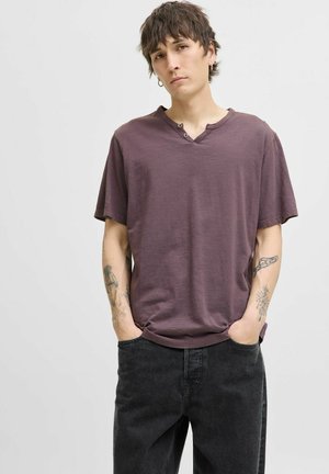 Camiseta de algodón morada de manga corta con cuello en V y dos detalles de botones. Ajuste relajado, textura suave, combinada con jeans oscuros.