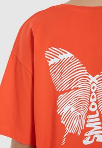 Oranžové bavlněné tričko s grafikou bílého motýla tvořenou otisky prstů na zádech. Volný střih, krátké rukávy a kulatý výstřih.