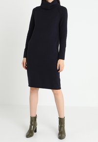 Robe ample bleu marine à col roulé et manches longues, en tissu doux, atteignant le milieu du mollet, assortie à des bottines vert émeraude brillantes.