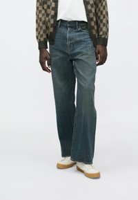 Jean en denim bleu foncé avec une coupe droite, présentant un léger effet délavé et un design classique à cinq poches, porté avec des baskets blanches.