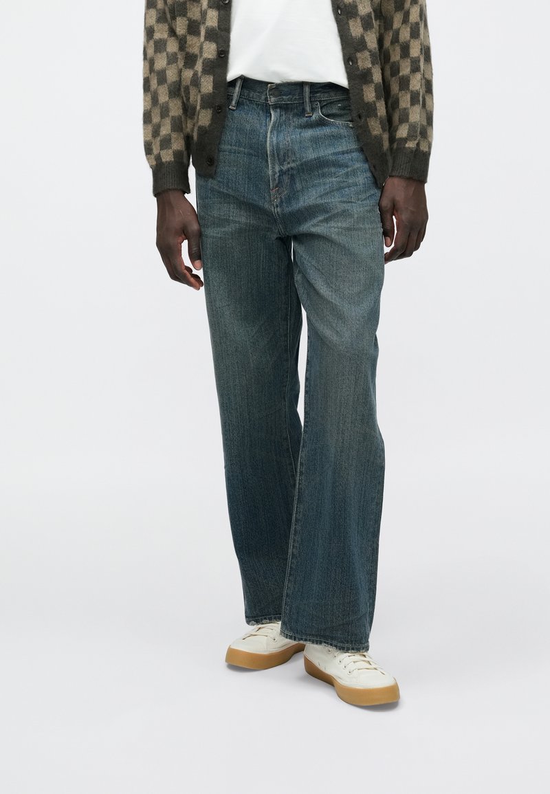 Jean en denim bleu foncé avec une coupe droite, présentant un léger effet délavé et un design classique à cinq poches, porté avec des baskets blanches.