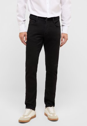 Mężczyzna w czarnych, dopasowanych dżinsach, białej koszuli z długim rękawem i beżowych trampkach stoi na tle jednolitego tła.