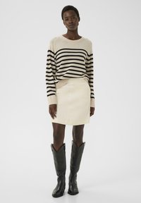 Pull rayé beige et noir, jupe en velours côtelé crème, et bottes de cow-boy noires à bouts pointus, offrant une silhouette structurée.