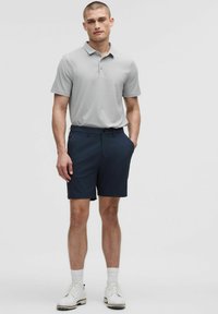 Grijze polo met korte mouwen en drie knopen, gecombineerd met marineblauwe shorts. Model draagt witte schoenen en sokken en staat met zijn handen in zijn zakken.