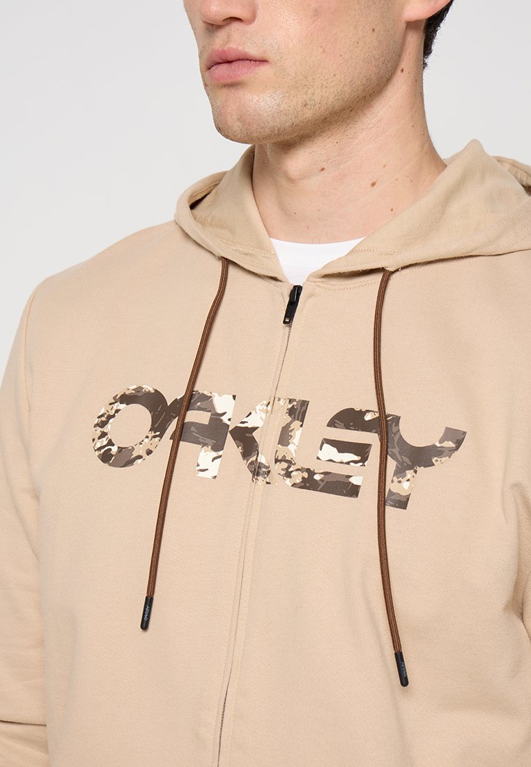 Sweatshirt à fermeture éclair beige en matériau doux, avec un logo camouflage brun foncé, des cordons de serrage marron et une coupe décontractée.
