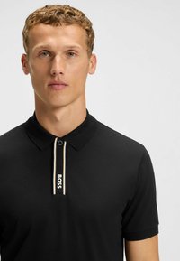 Black short-sleeve polo shirt with a button-up collar, featuring a white and cream trim and embroidered "BOSS" logo on the placket.