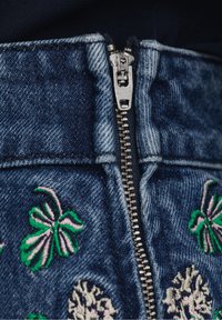 Prenda de mezclilla con un zipper metálico plateado, que presenta bordados florales en verde y rosa en la cinturilla. Tela gruesa y texturizada.