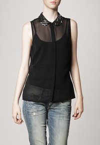 Blusa nera senza maniche in tessuto trasparente con colletto geometrico di perline. Presenta chiusura frontale con bottoni e tasche laterali con texture in contrasto.