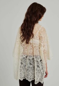 Sovrapposizione in pizzo stile kimono, con intricati motivi floreali, tessuto crema semi-trasparente e una silhouette fluttuante, indossata sopra abbigliamento scuro.