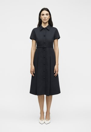 Kvinde stående iført en sort midi-kjole med knapper, korte ærmer, krave, bælte og hvide spidse sko på en ensfarvet hvid baggrund.