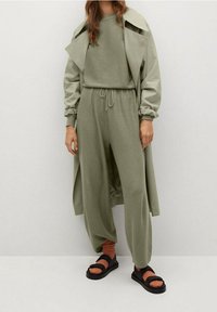 Olijfgroen outfit met een cropped sweatshirt en losse broek, gelaagd met een bijpassende trenchcoat. Draagt zwarte sandalen en oranje sokken.