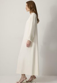 Witte maxi-jurk met lange, bolle mouwen en een rechte silhoutte. Gladde, lichte stof met een minimalistisch ontwerp. Gedragen met beige hakken.