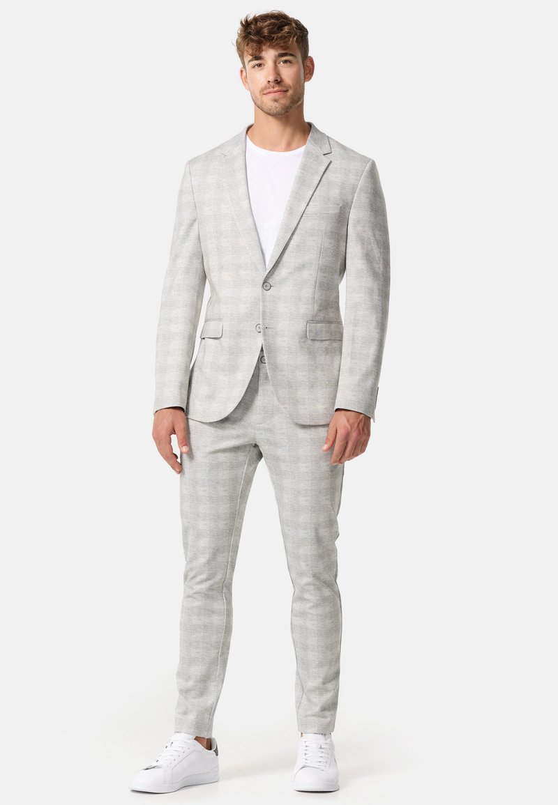 Costum în carouri gri deschis, realizat dintr-un material texturat, cu un sacou cu două butoane și pantaloni tapered, asortat cu un tricou alb și adidași albi.