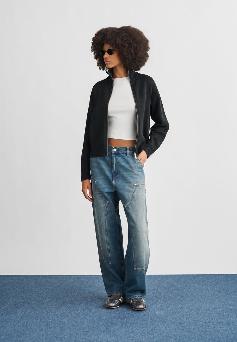 Black zip-up jacket, white cropped top, and loose blue jeans with visible seams. Gray footwear. Background is plain and light-colored.