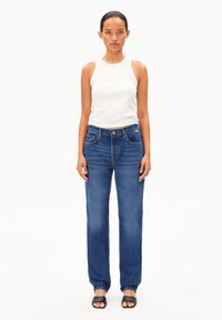 Débardeur blanc léger à côtes associé à un jean droit bleu foncé avec cinq poches et un aspect légèrement délavé. Sandales noires à enfiler.