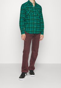 Camisa de cuadros en verde y negro con dos bolsillos en el pecho, combinada con jeans burdeos y zapatillas negras con un logo blanco.