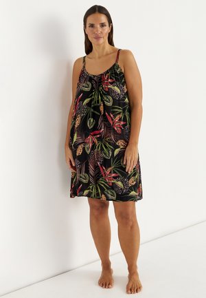 Kvinde stående barfodet iført en ærmeløs, knælange sort kjole med grønne og røde tropiske bladmønstre.