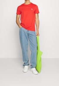 Camiseta roja de manga corta con un pequeño logo, combinada con jeans de mezclilla azul claro y zapatillas blancas. Sosteniendo una bolsa de mano verde brillante.