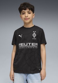 Ragazzo che indossa una maglia da calcio nera del Borussia Mönchengladbach con loghi bianchi e jeans azzurri chiari su uno sfondo grigio.