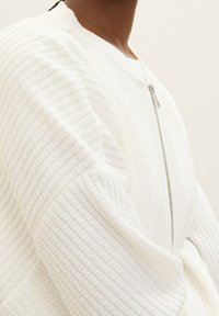 Giacca bianca con zip in tessuto strutturato e rigato. Presenta un collo rotondo e maniche ampie, mettendo in risalto un design sobrio e strutturato.