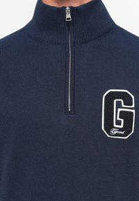Navy blue knit sweater with a half zipper front and a large black and white "G" patch on the left chest.